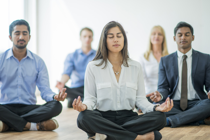 A multi-ethnic group of young business men and women in semi-casual office clothes are sitting on the floor and meditating to relax in an indoor, sunlit office.