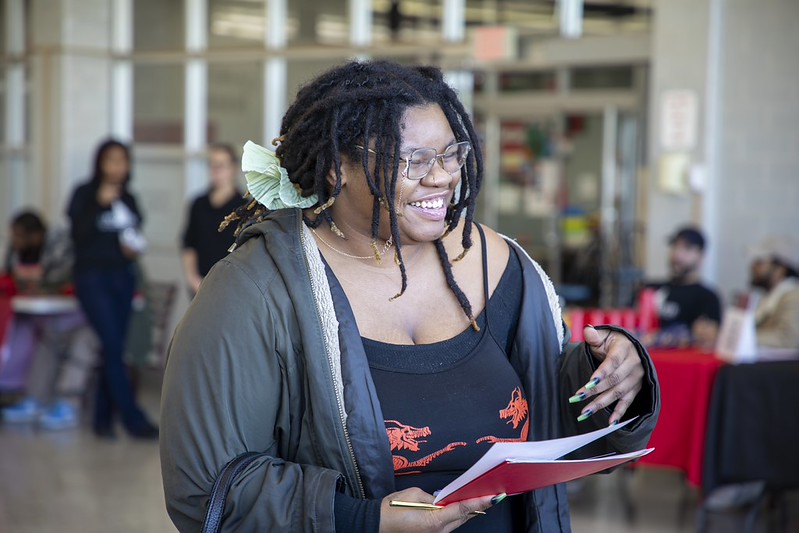 A student smiles while holding papers and speaking with someone during an event in the north campus cafeteria at SUNY Erie.