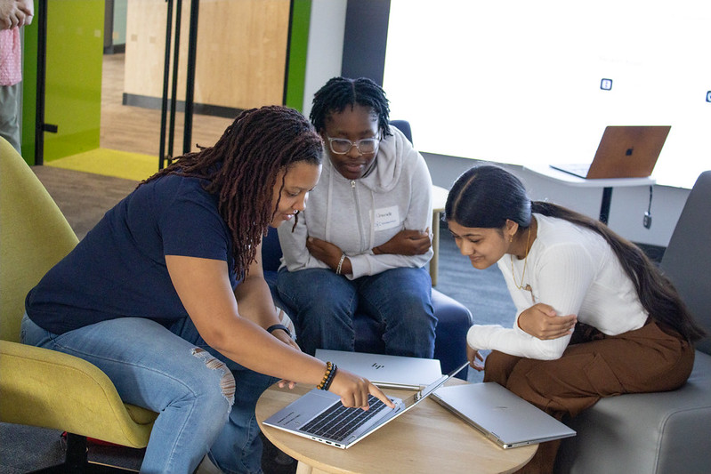 Three girls sit together in a modern lounge space, gathered around an open laptop. The girl on the left points to the screen while the other two lean in closely, smiling and engaged in the discussion. Additional laptops and seating are visible in the background.