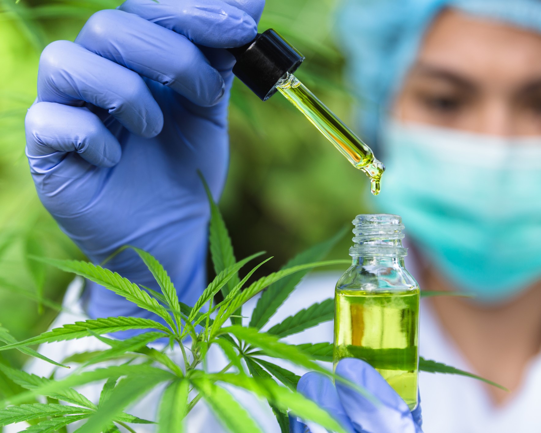 Woman with hairnet & facemask holding a small vial of green oil in her left hand, and a dropper in her right.